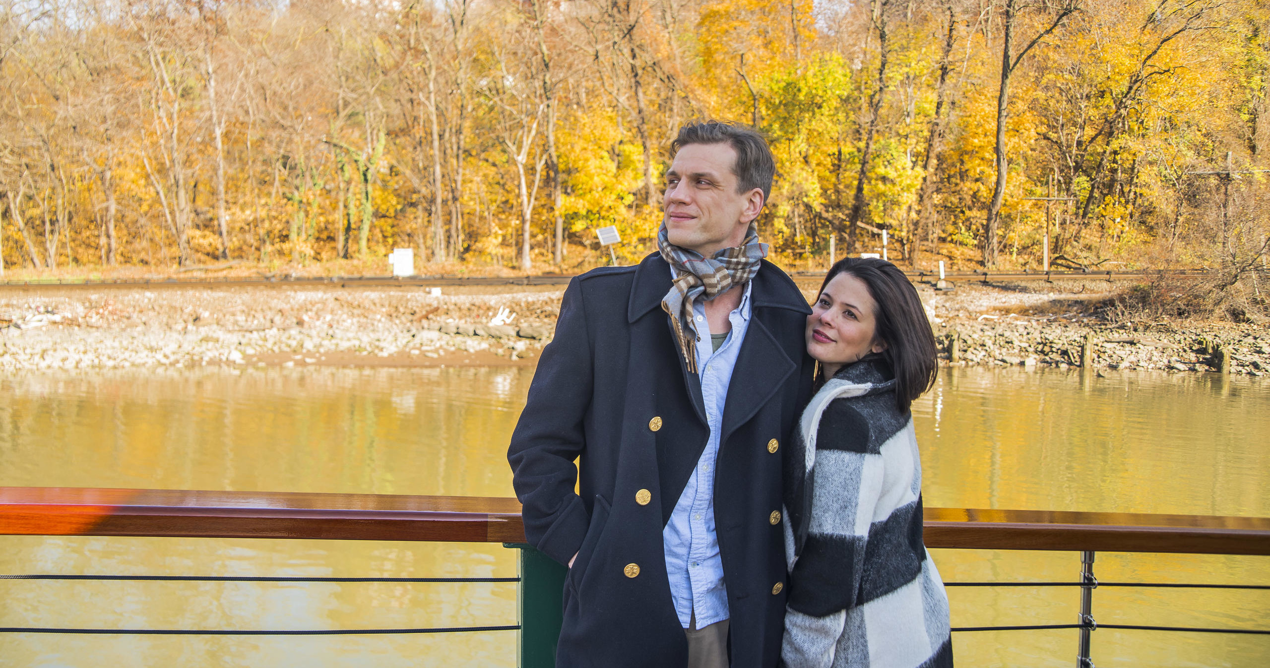 Couple stands by river, autumn trees in background.