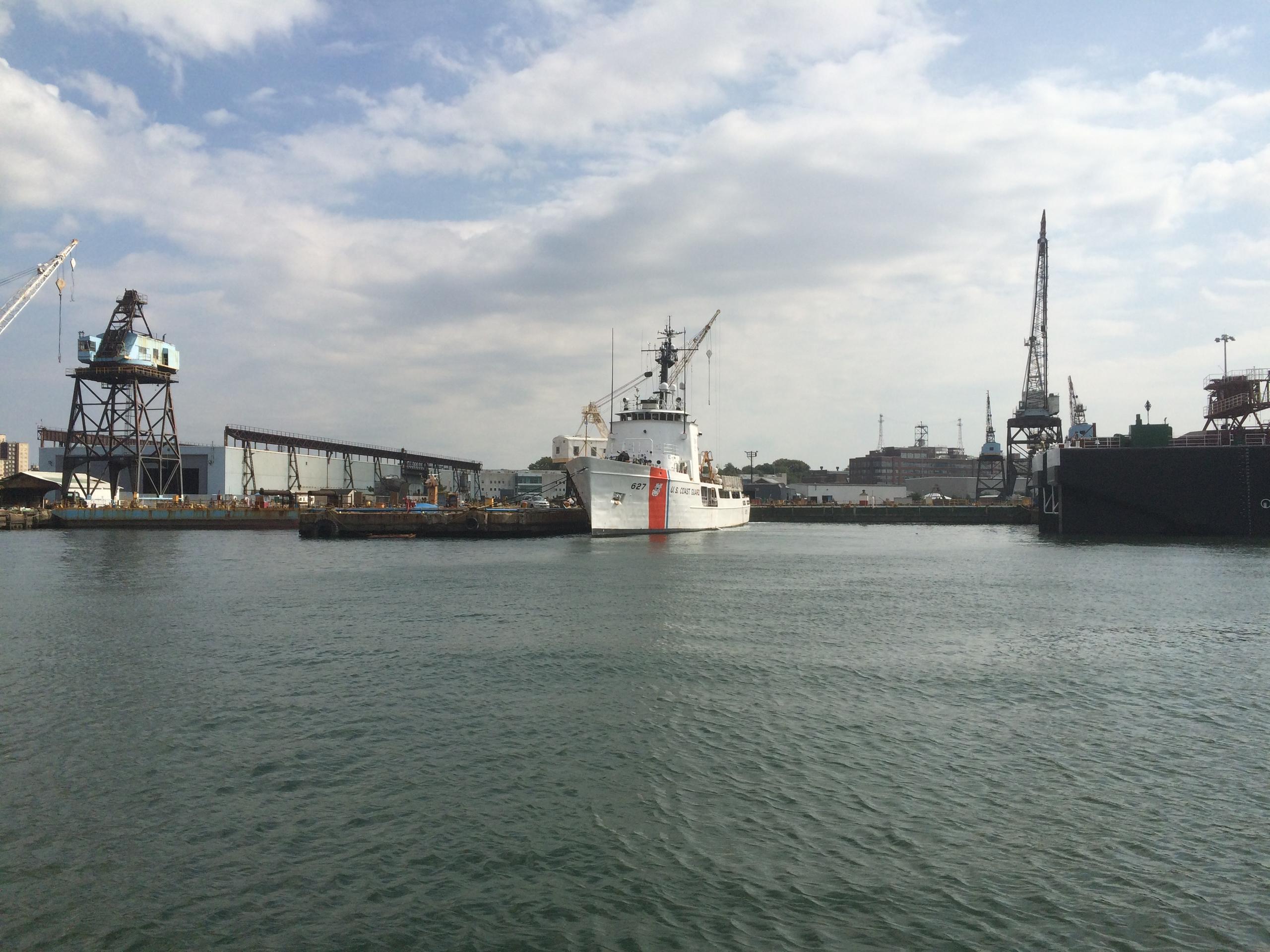 Ship docked at an industrial port with cranes and warehouses under a cloudy sky.