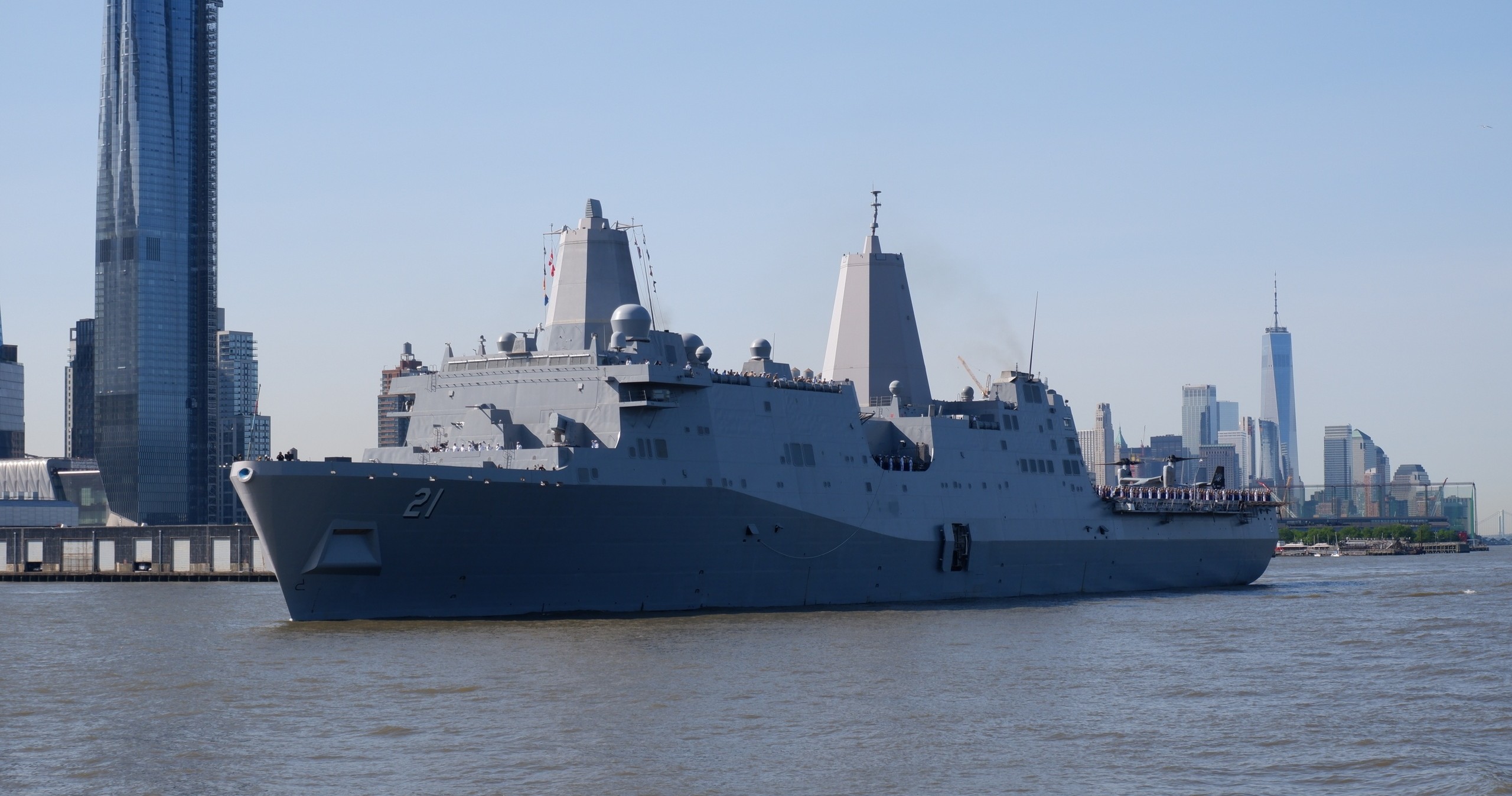 Large navy ship sailing near skyscrapers in a city skyline.
