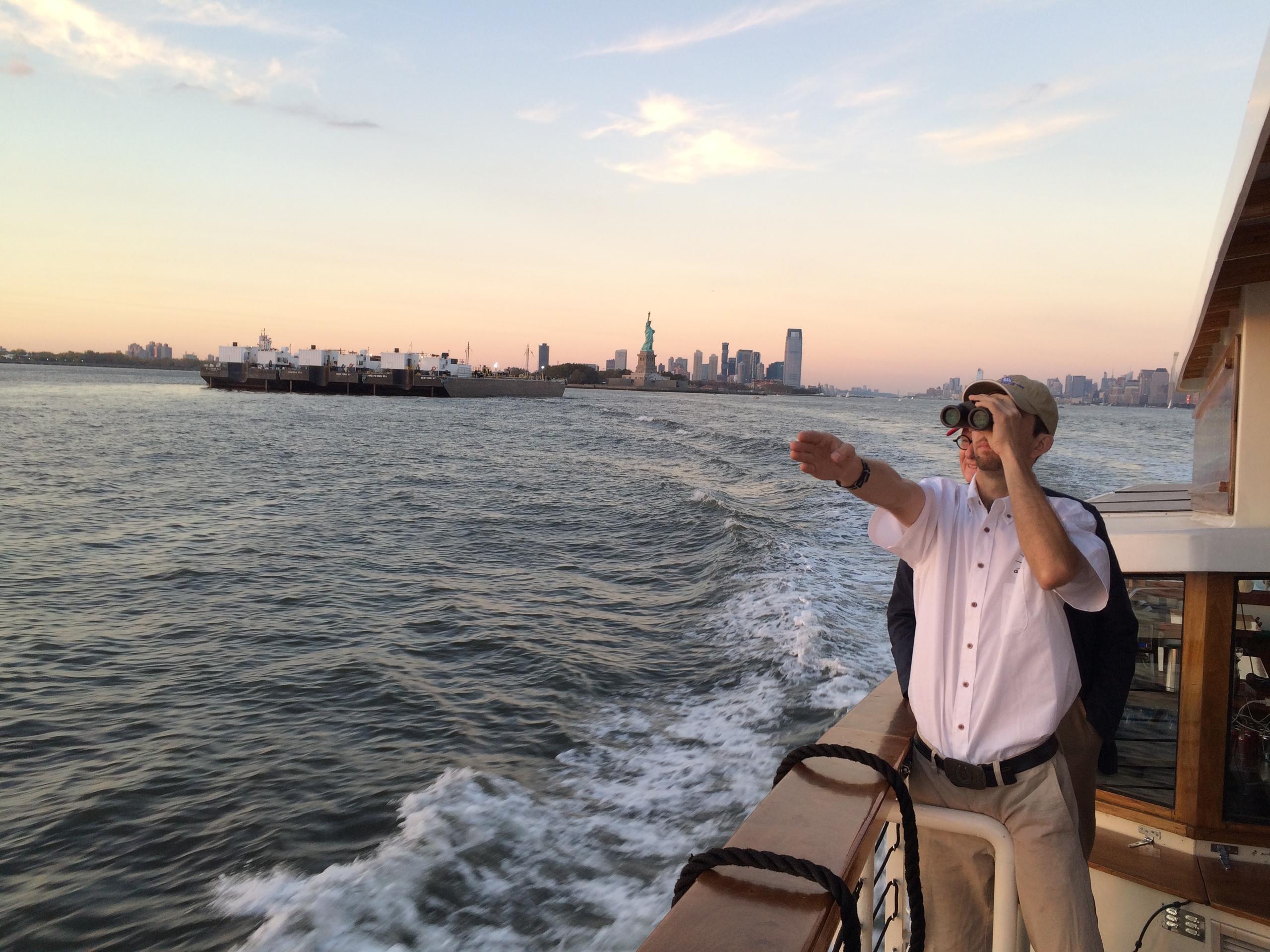Person on boat pointing towards distant Statue of Liberty at sunset.