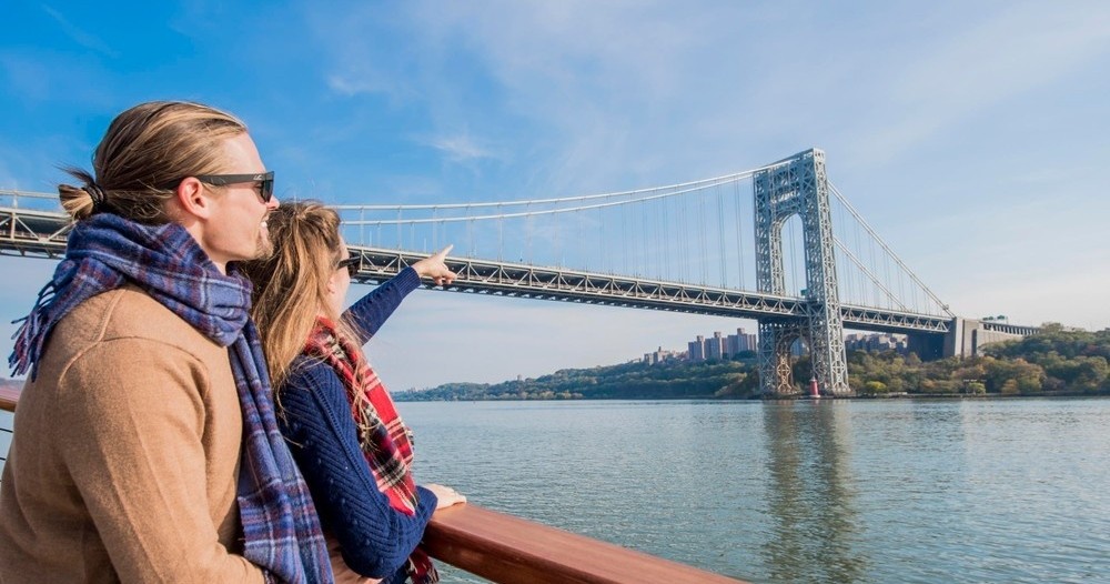 Two people on a boat looking at a large suspension bridge over calm water.