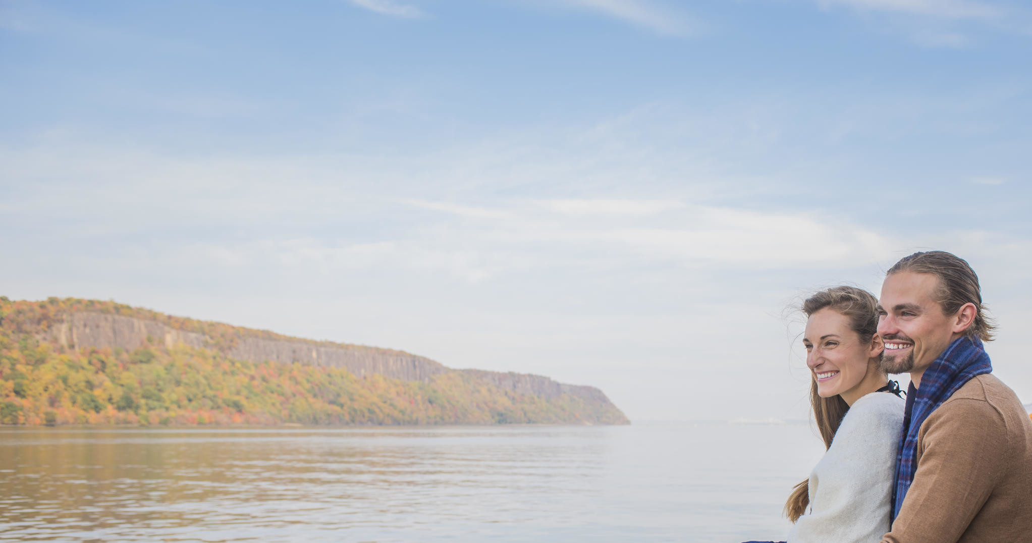 Smiling couple sitting by a calm lake with cliffs in the background.