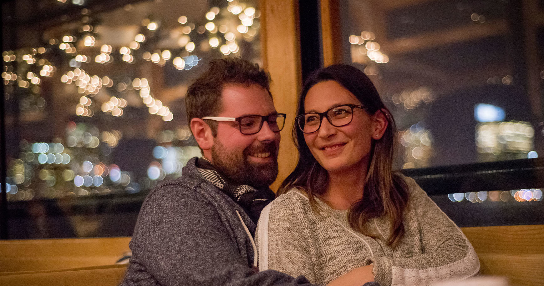 Smiling couple with glasses sitting together inside a warmly lit room.