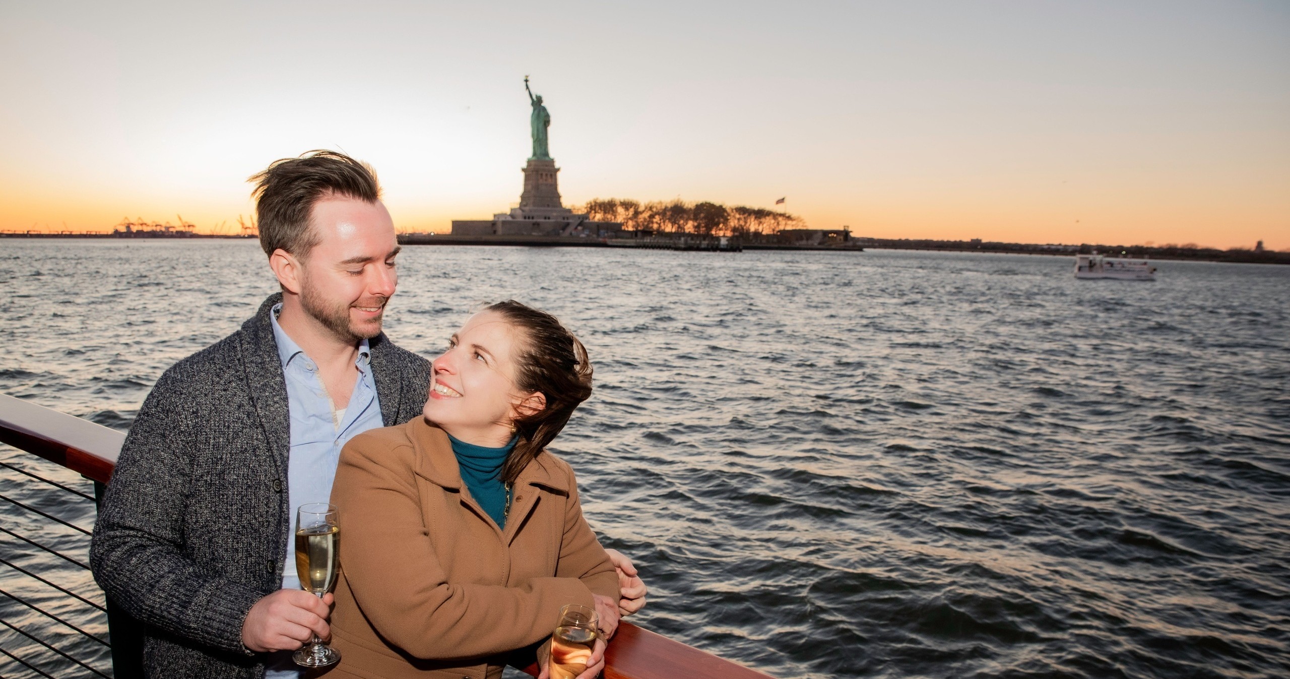 Couple on a boat with drinks, Statue of Liberty in the background at sunset.