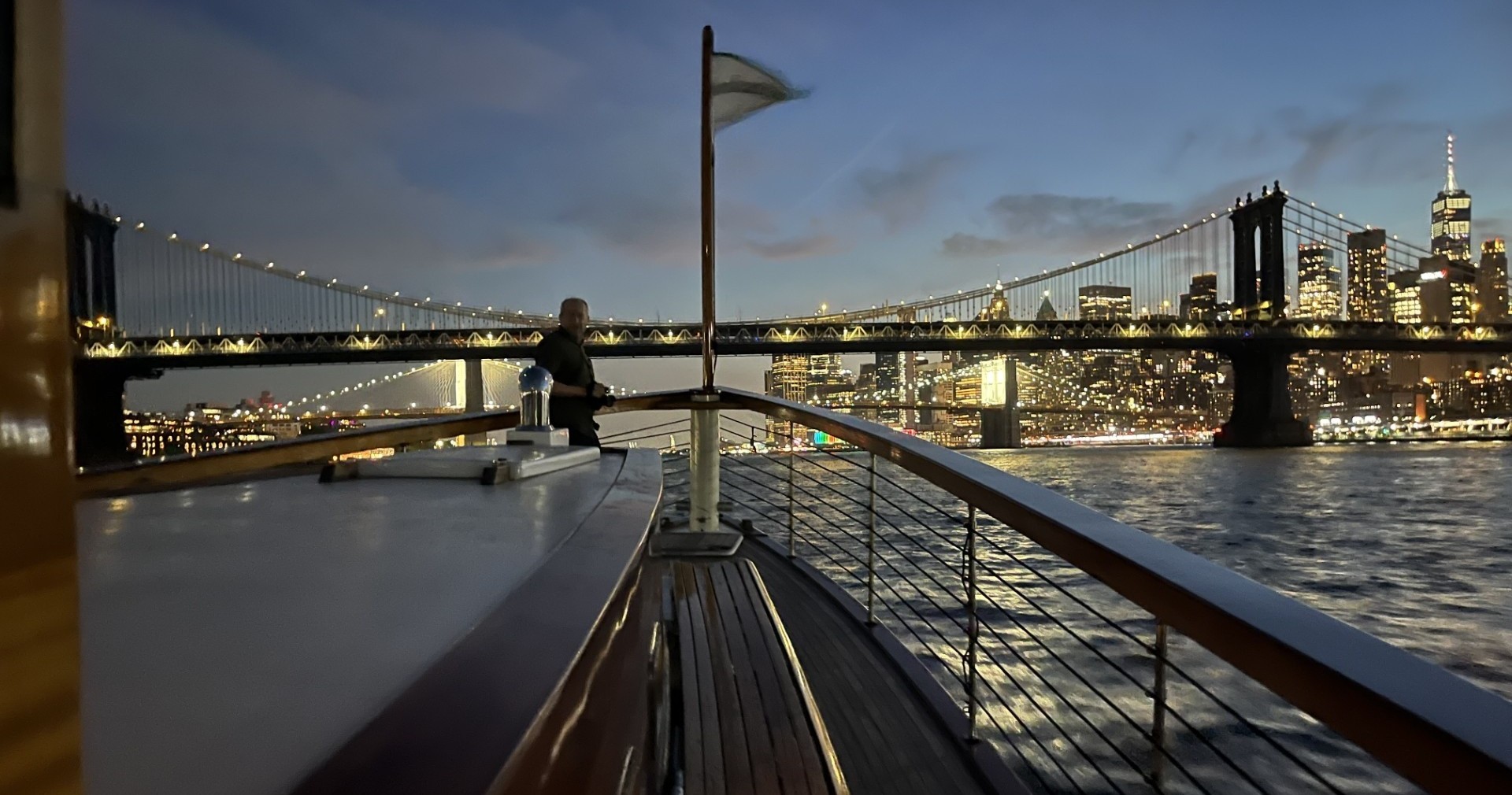 Boat on a river at dusk with a lit bridge and city skyline in the background.
