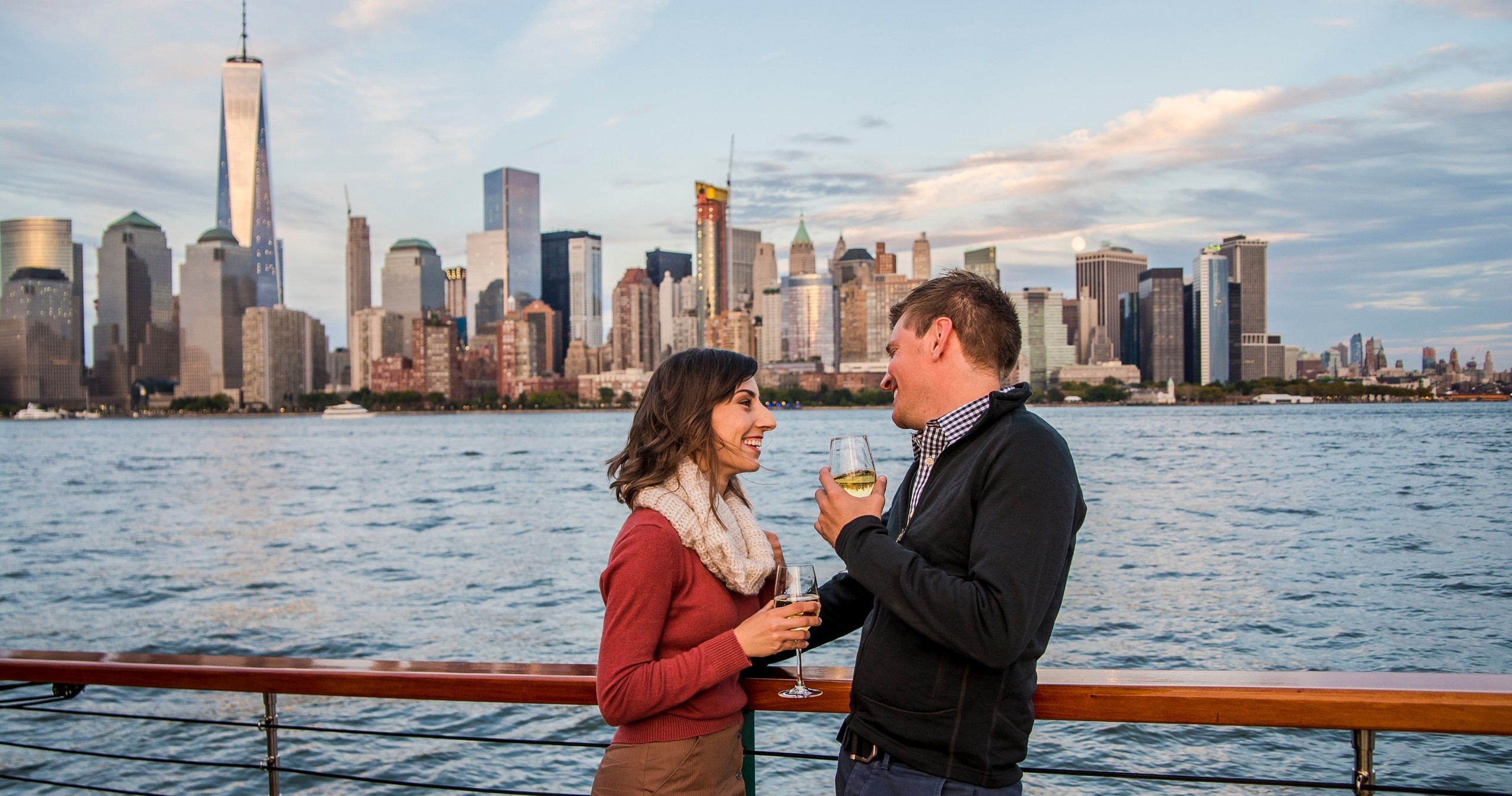 Couple on a boat with drinks, New York City skyline in background.