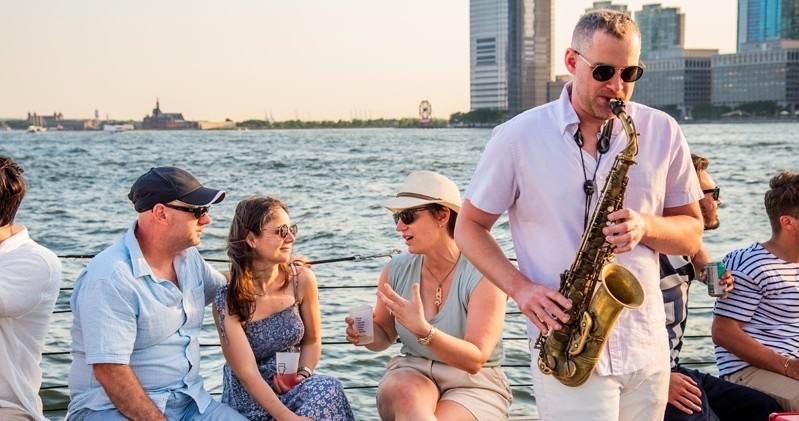 People enjoy music by a saxophonist near a waterfront, city buildings in the background.