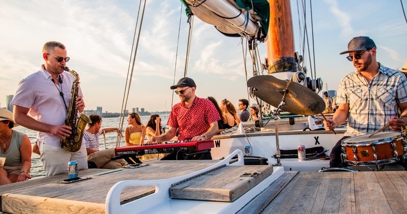 Musicians play saxophone, keyboard, and drums on a boat with seated people enjoying the scene.