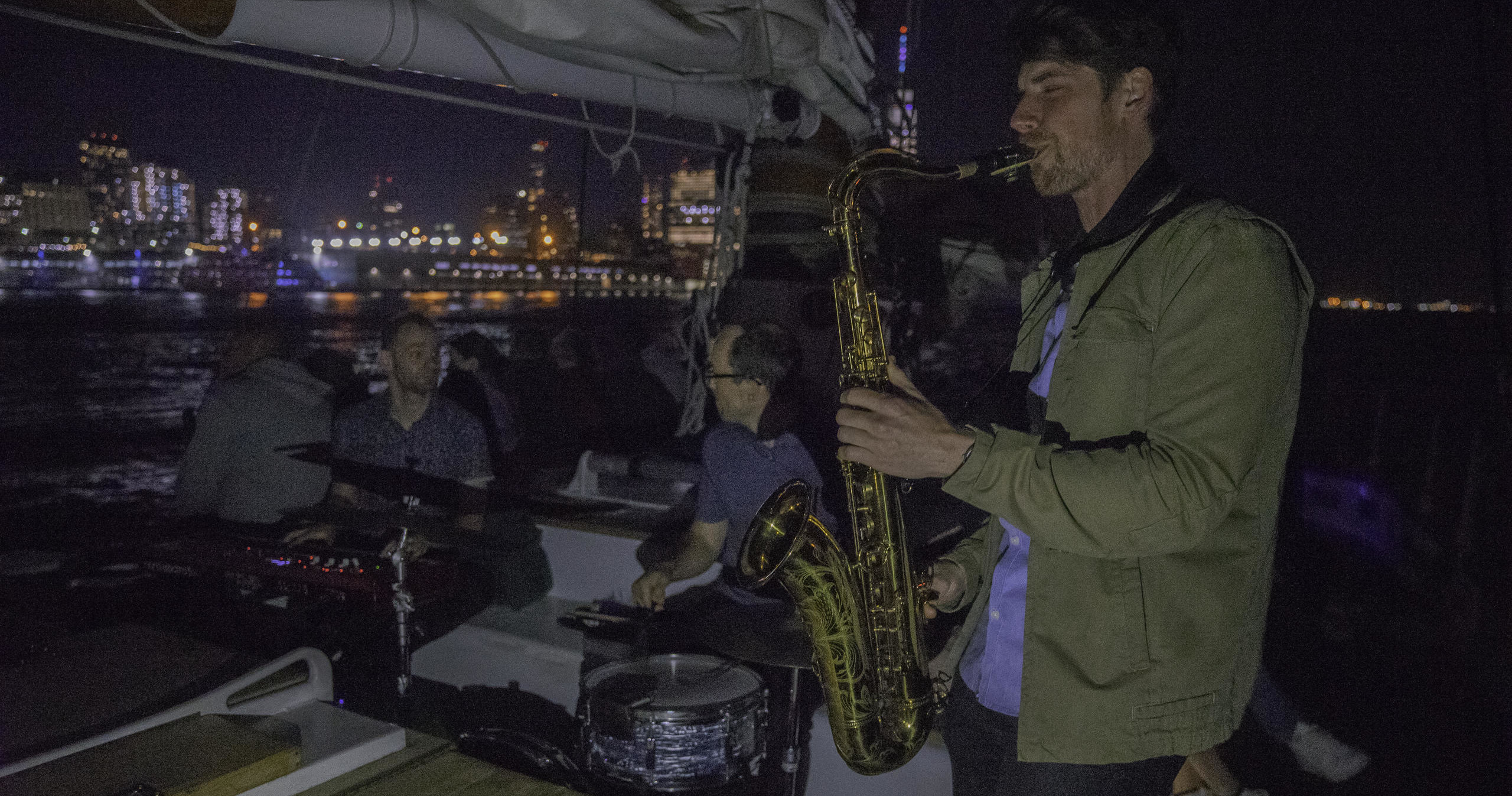 Saxophonist plays on a boat at night with city skyline in the background.