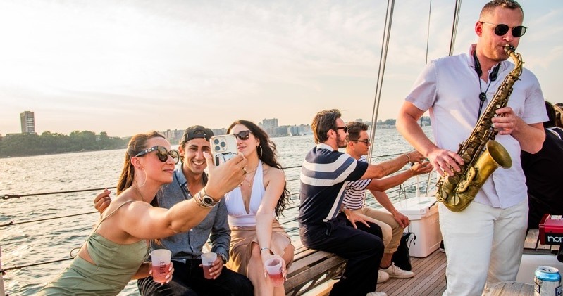 Group on a boat with drinks, one playing saxophone, cityscape in background.