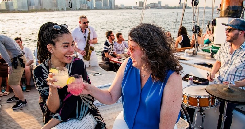 Two women laughing and clinking drinks on a boat with people in the background.