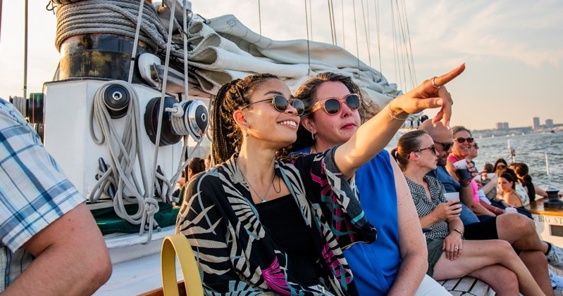Two women sitting on a boat, one pointing, with sails and water in the background.