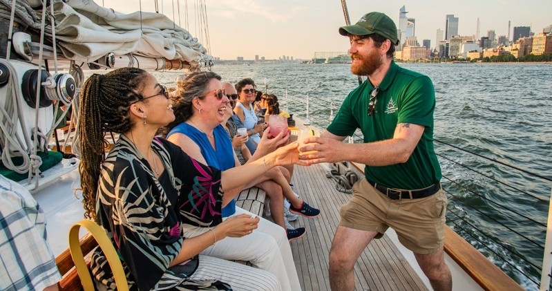 Smiling crew member serves drinks to seated women on a sailing boat.