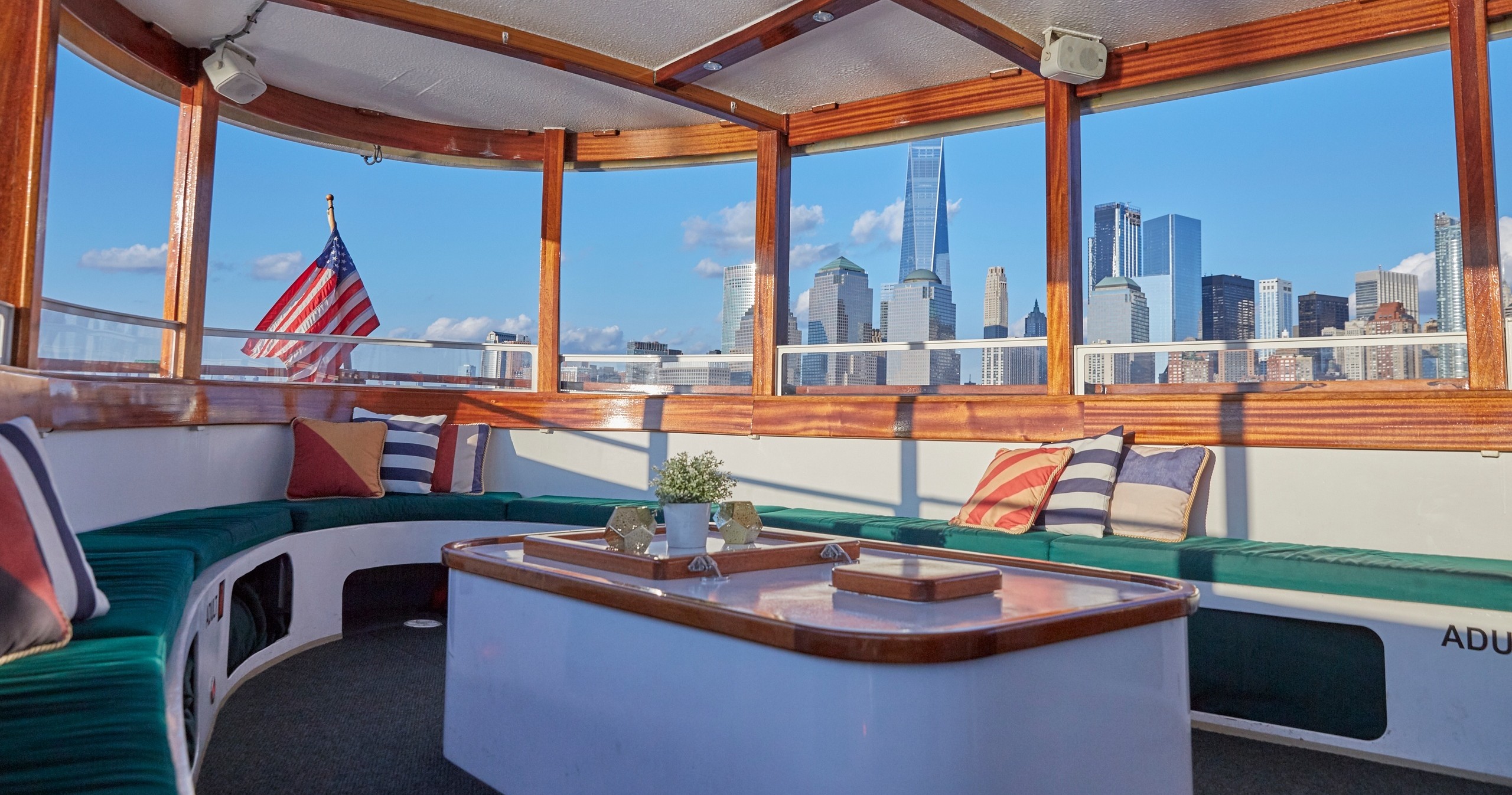 Boat interior with city skyline view, American flag, green cushions, and a central white table.