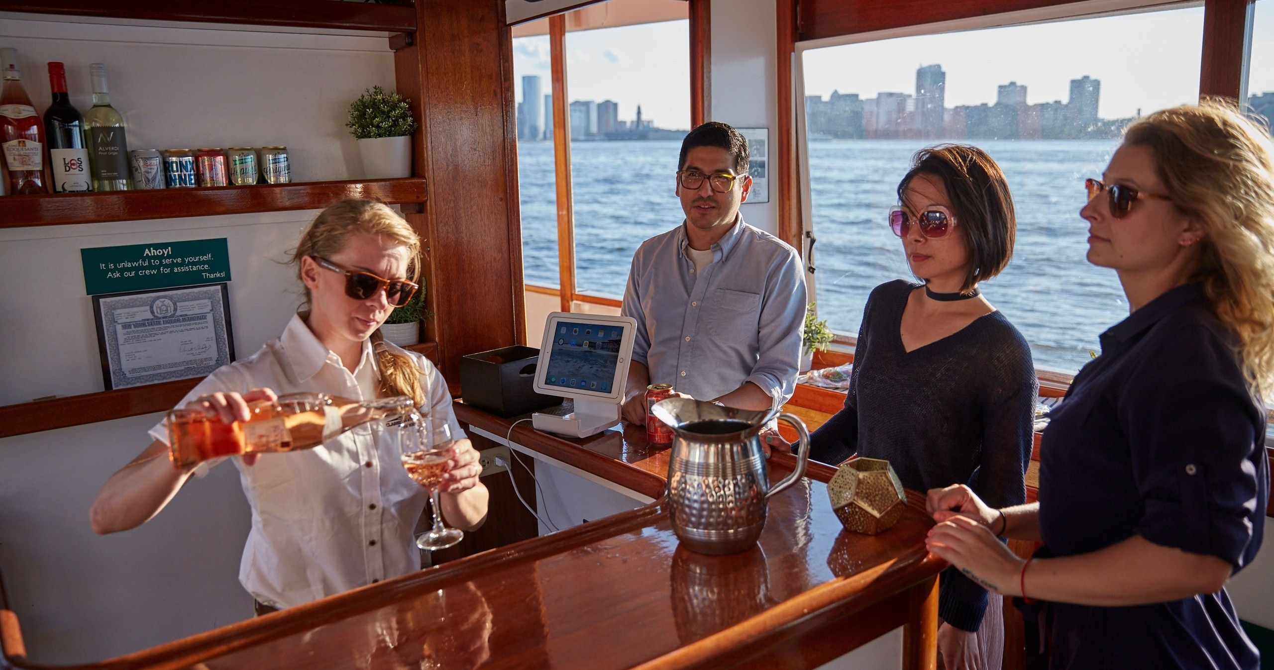People in sunglasses at a boat bar with a cityscape in the background.