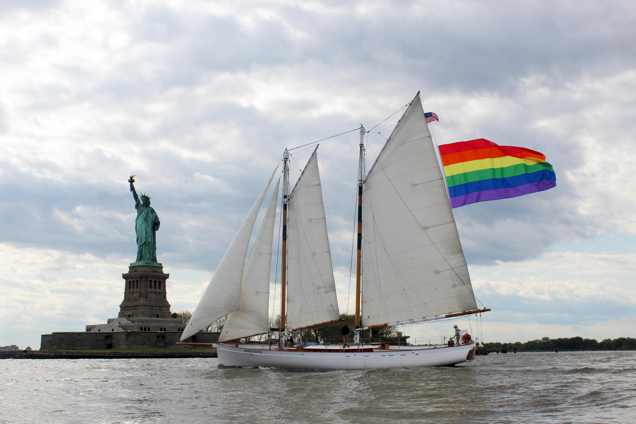 Sailboat with rainbow flag near the Statue of Liberty on a cloudy day.