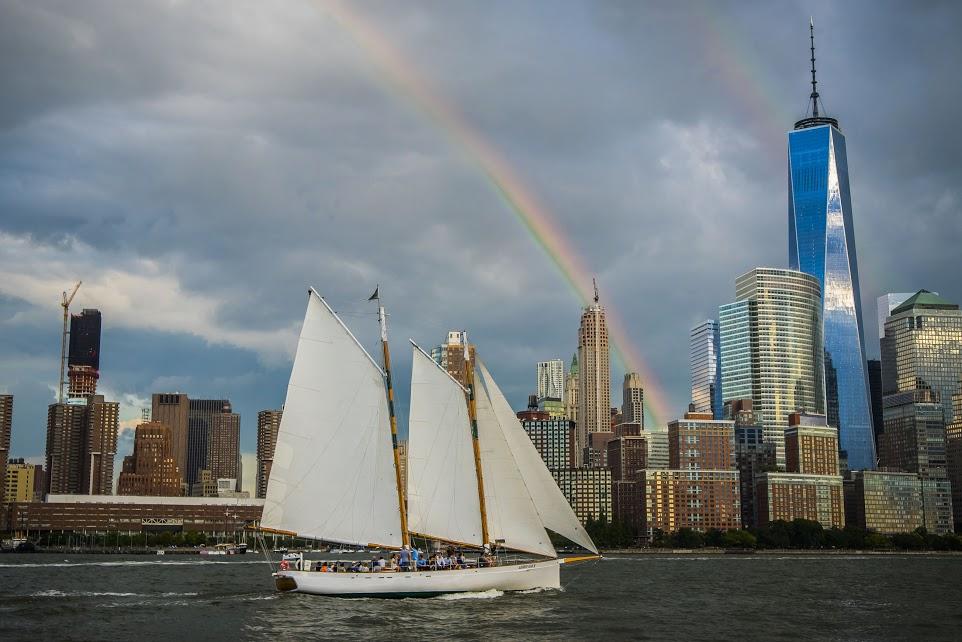 Sailboat on river with city skyline and rainbow in background.