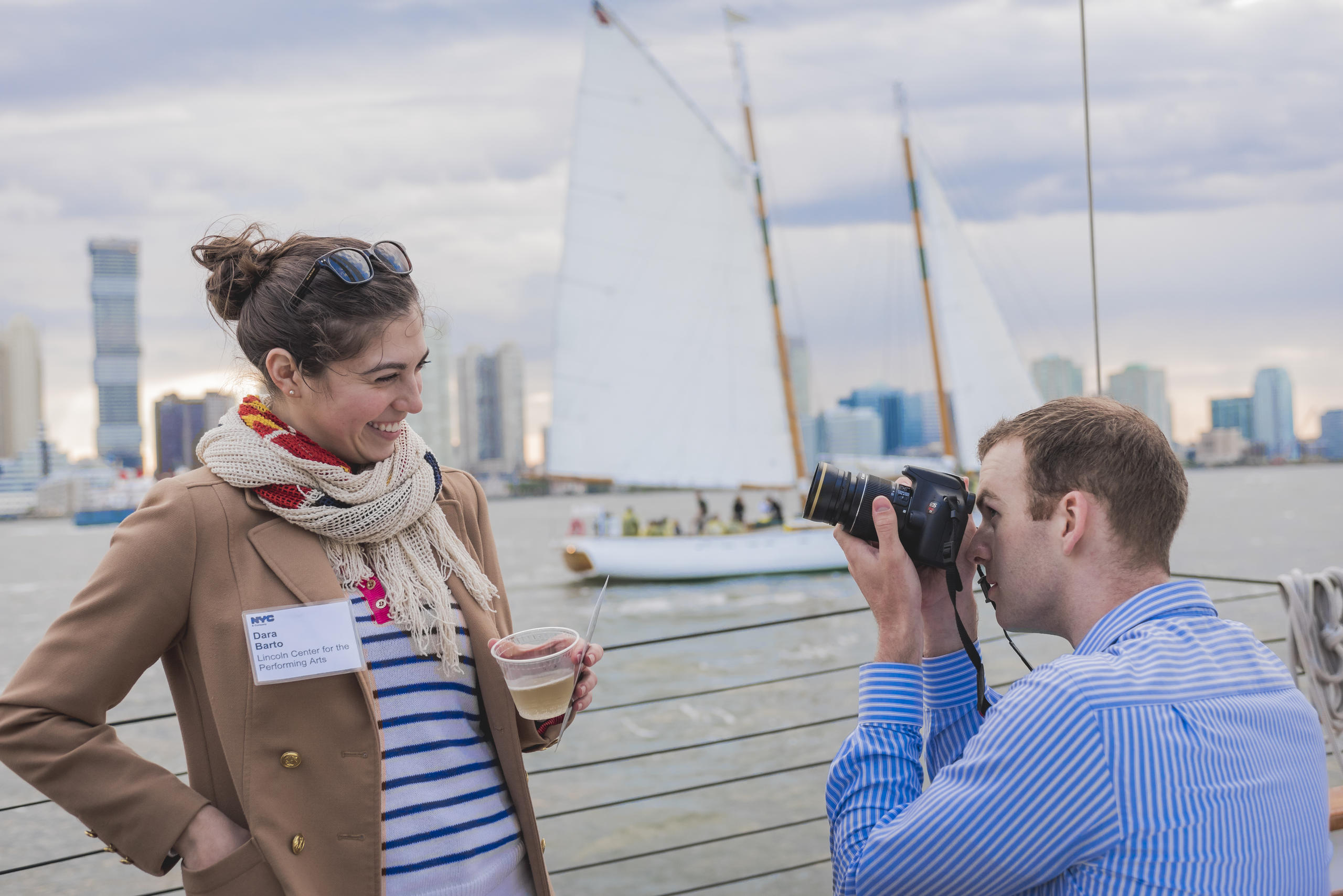 Woman poses with drink on a boat while man takes her photo, cityscape and sailboat in background.