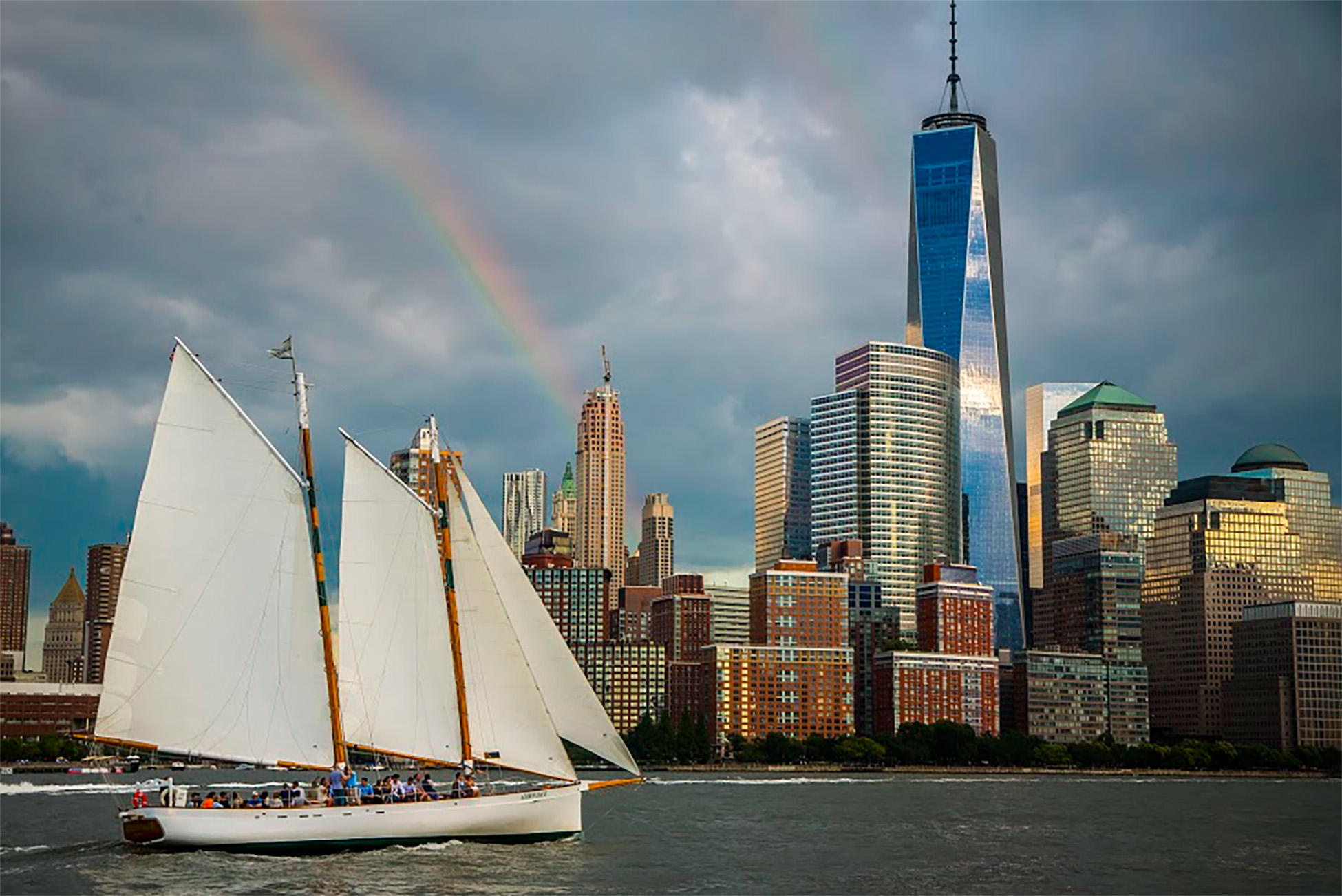 Sailboat on water with a city skyline and a rainbow in the cloudy sky.