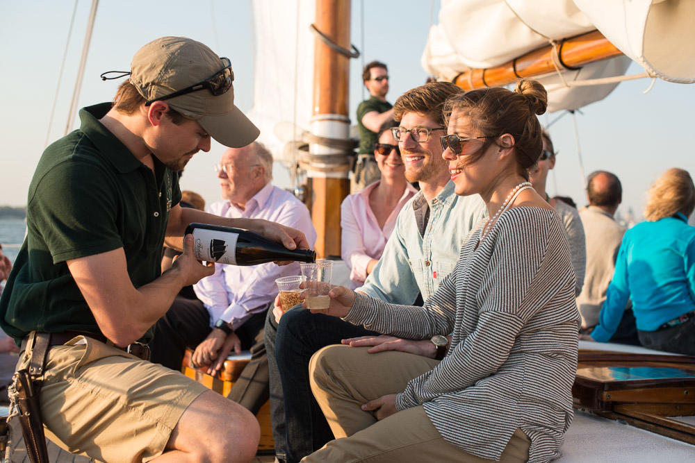 People on a boat enjoying drinks, with a man pouring wine for a seated couple.