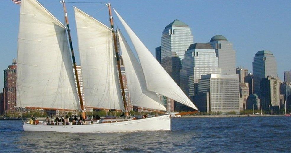 Sailboat with white sails near modern skyscrapers on a sunny day.