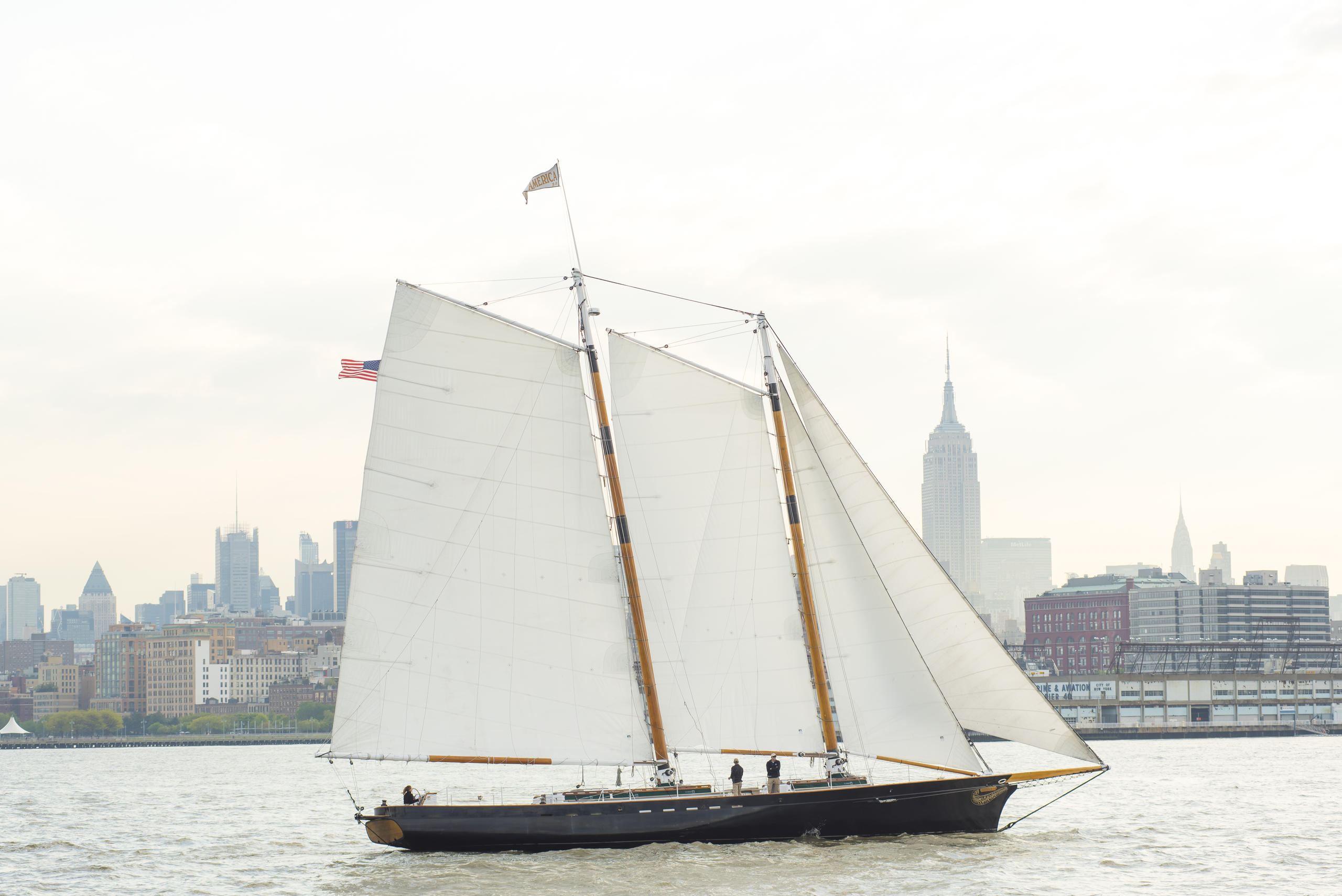 Sailboat with white sails in front of city skyline on cloudy day.