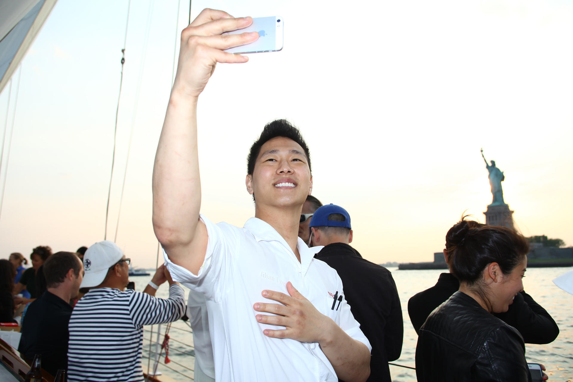 Man taking a selfie on a boat near the Statue of Liberty with a group of people.