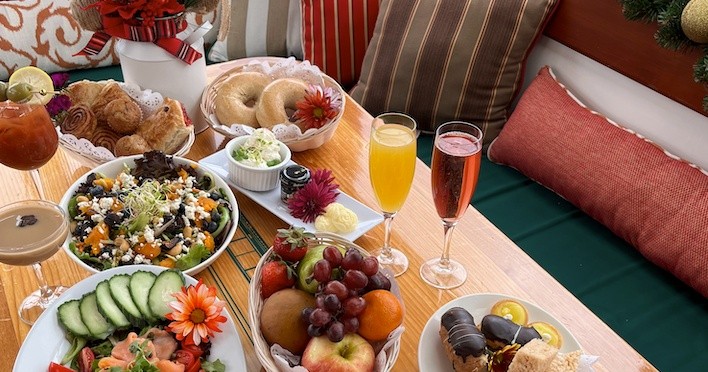 Colorful brunch spread with salads, bagels, fruit, and drinks on a table with cushions.