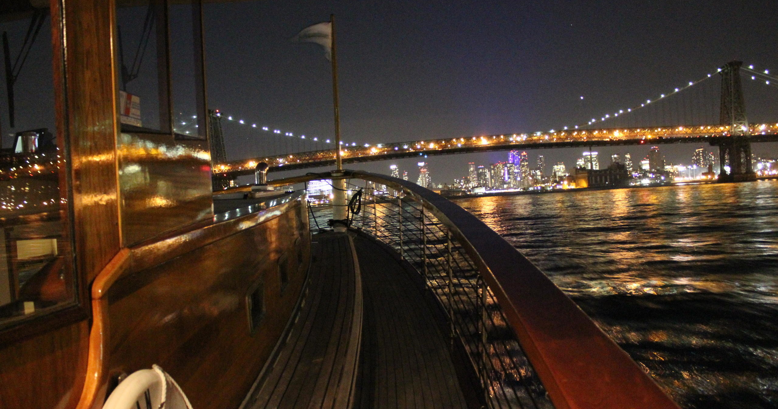 View from a boat at night, city skyline and lit bridge in the background.