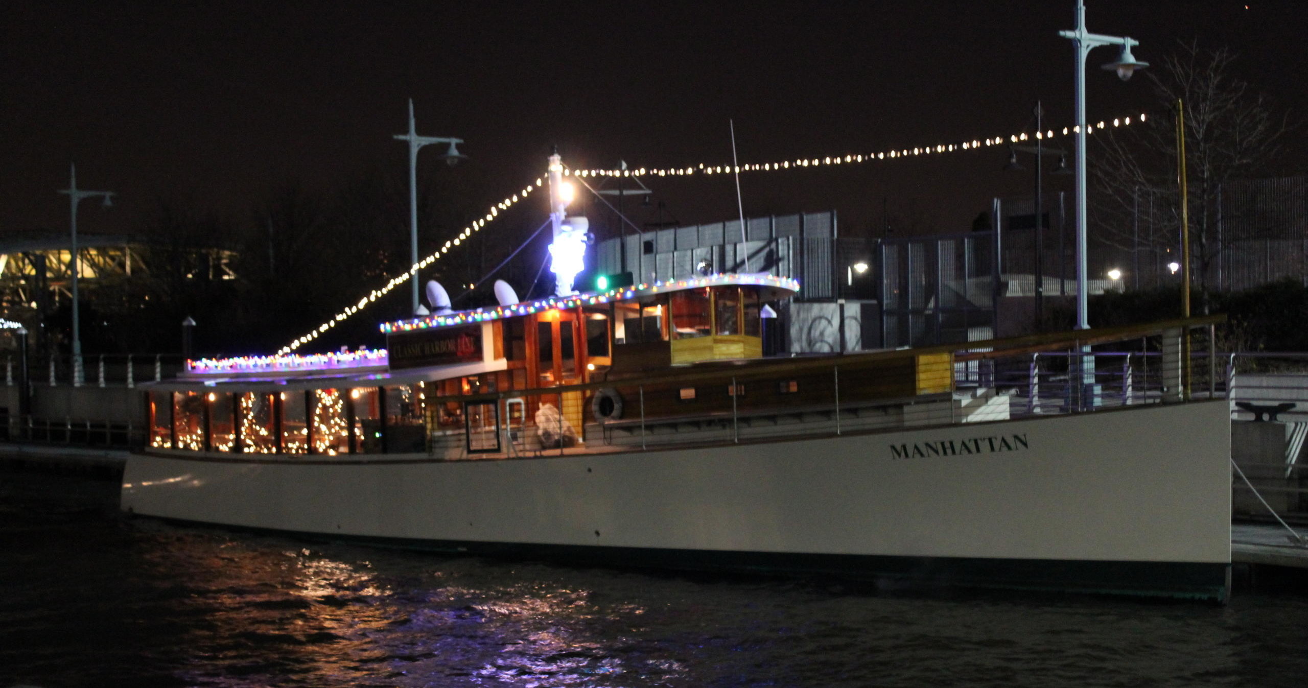 Illuminated boat named Manhattan at night, decorated with festive lights.