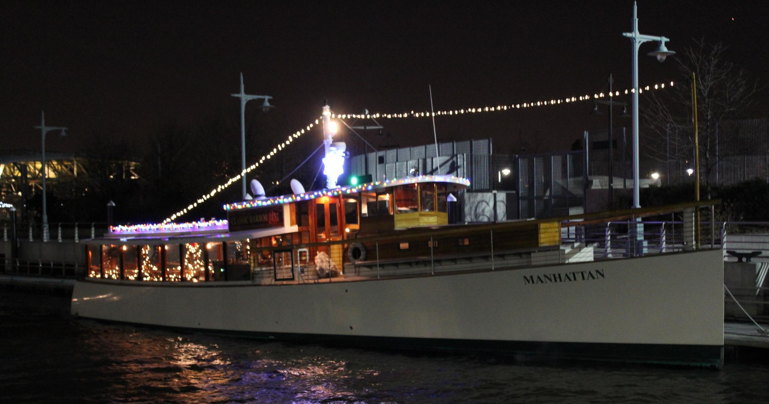 Boat named Manhattan adorned with string lights, docked at night.