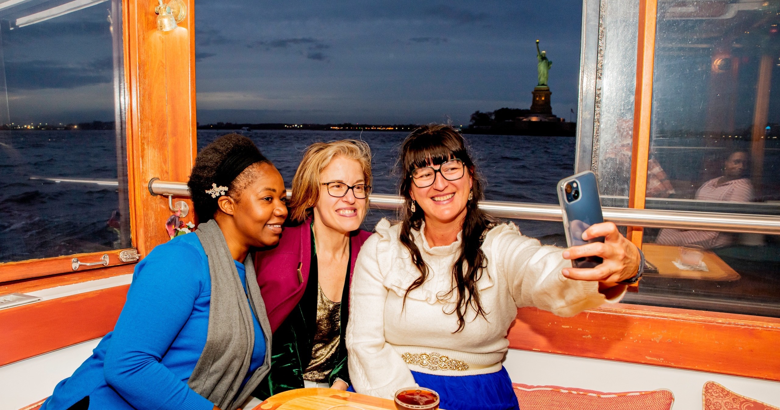Three women taking a selfie on a boat with the Statue of Liberty in the background.
