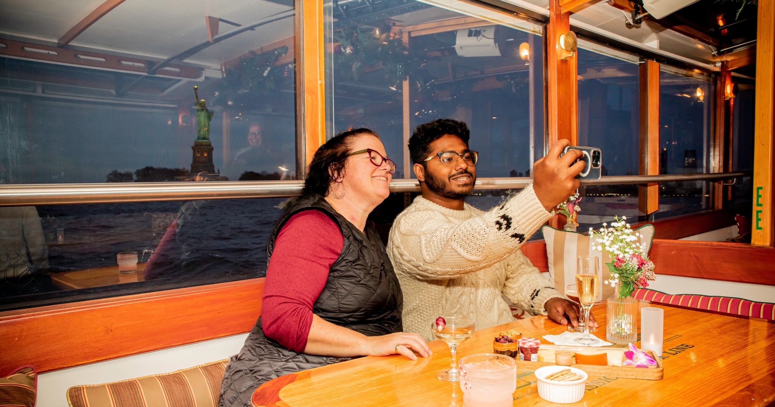 Two people take a selfie at a table on a boat with the Statue of Liberty in the background.