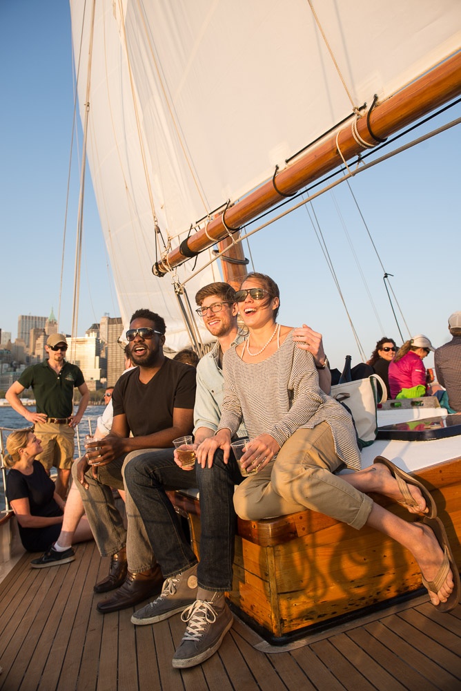 Three people smiling on a sailboat deck with city skyline in the background.