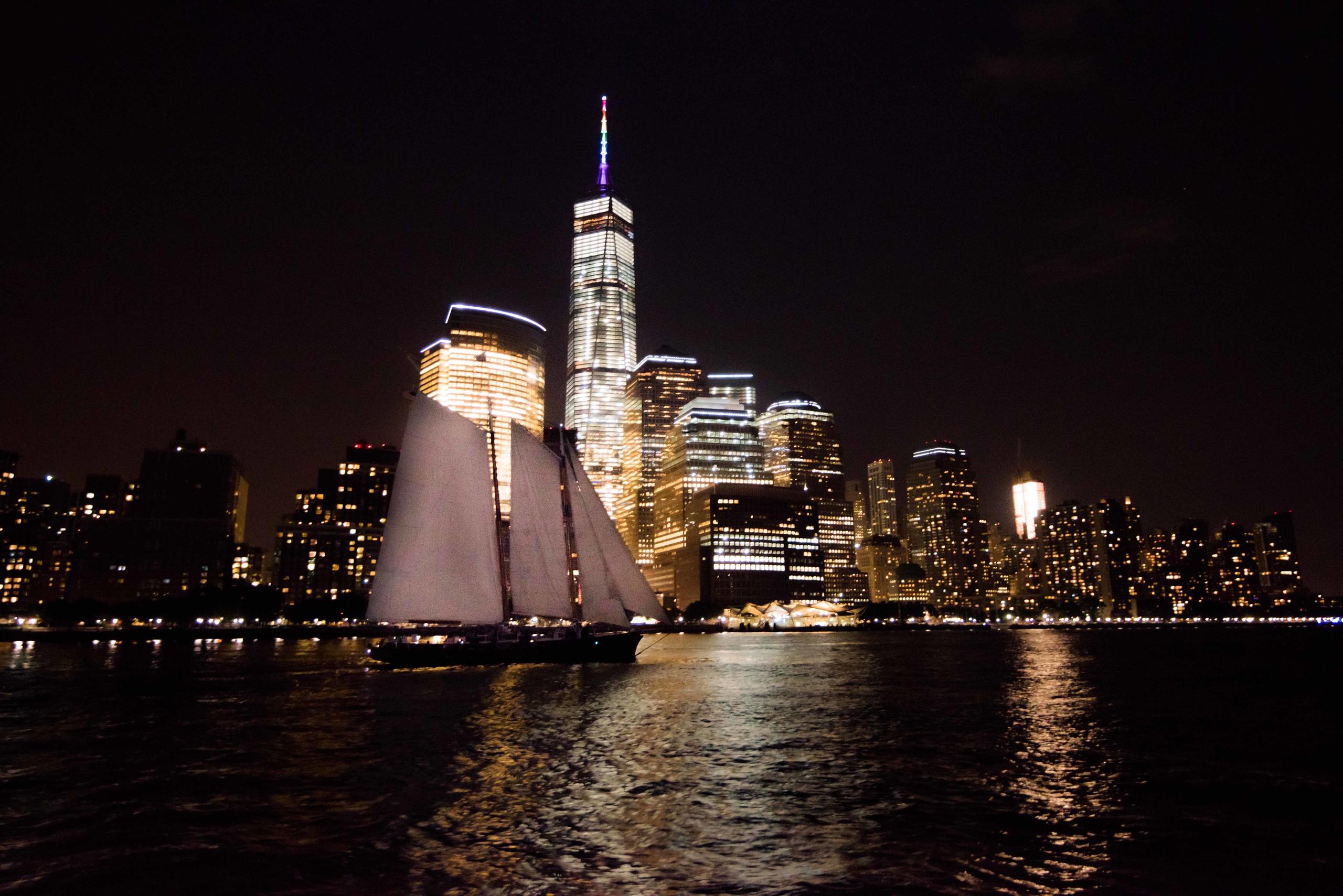 Sailboat on water at night with illuminated city skyline in background.