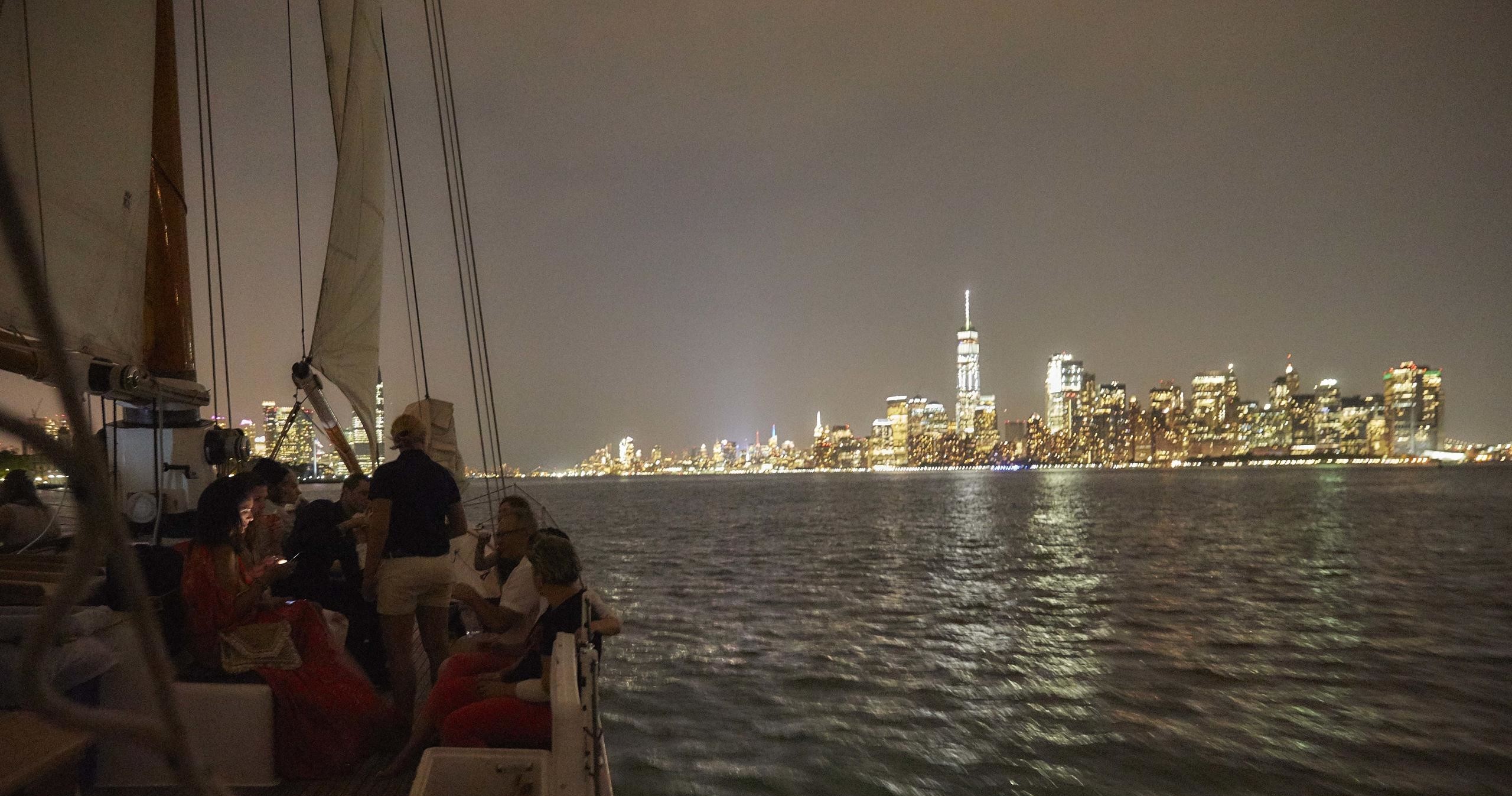 Sailboat with people at night near a city skyline with illuminated buildings.