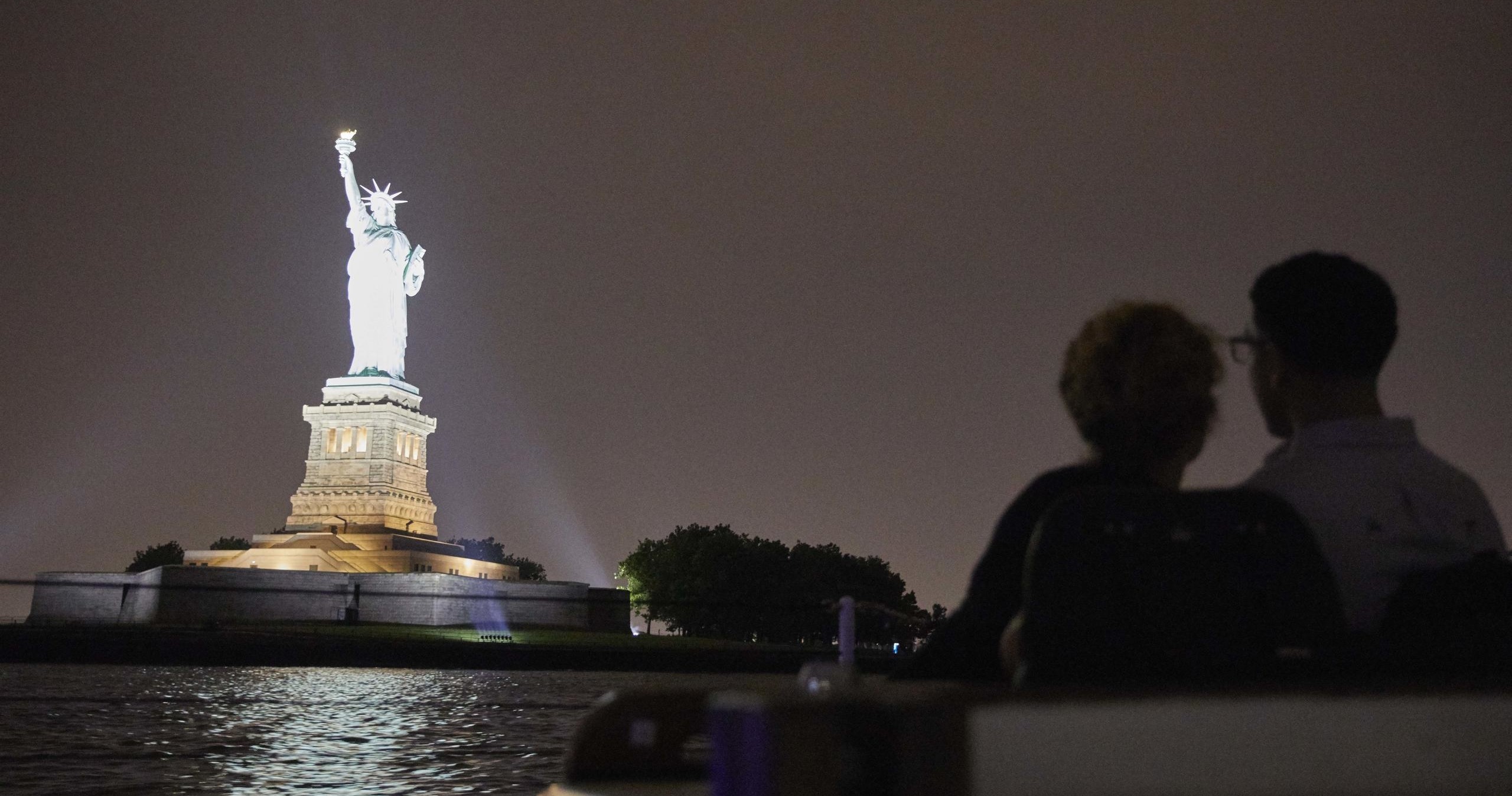Two people seen in silhouette view the illuminated Statue of Liberty at night.