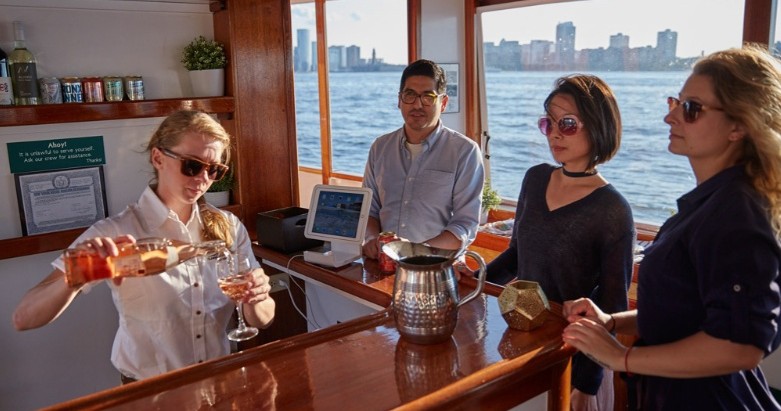 Bartender serving drinks to three people on a boat with a city skyline in the background.