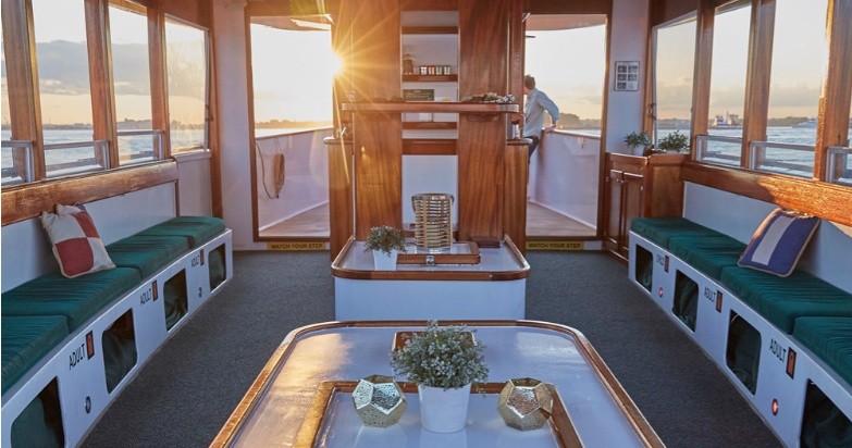 Boat interior with sunlit windows, green benches, and decorative plants.