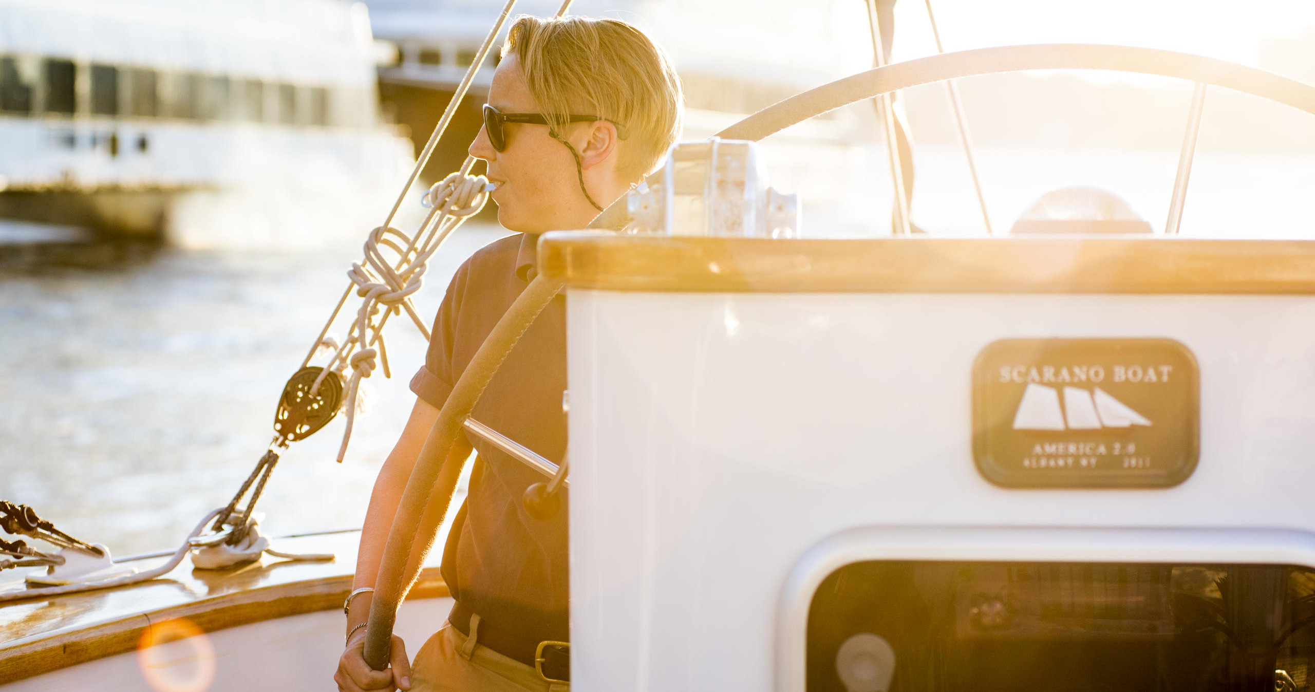 Person steering a boat in sunlight, wearing sunglasses and casual attire.