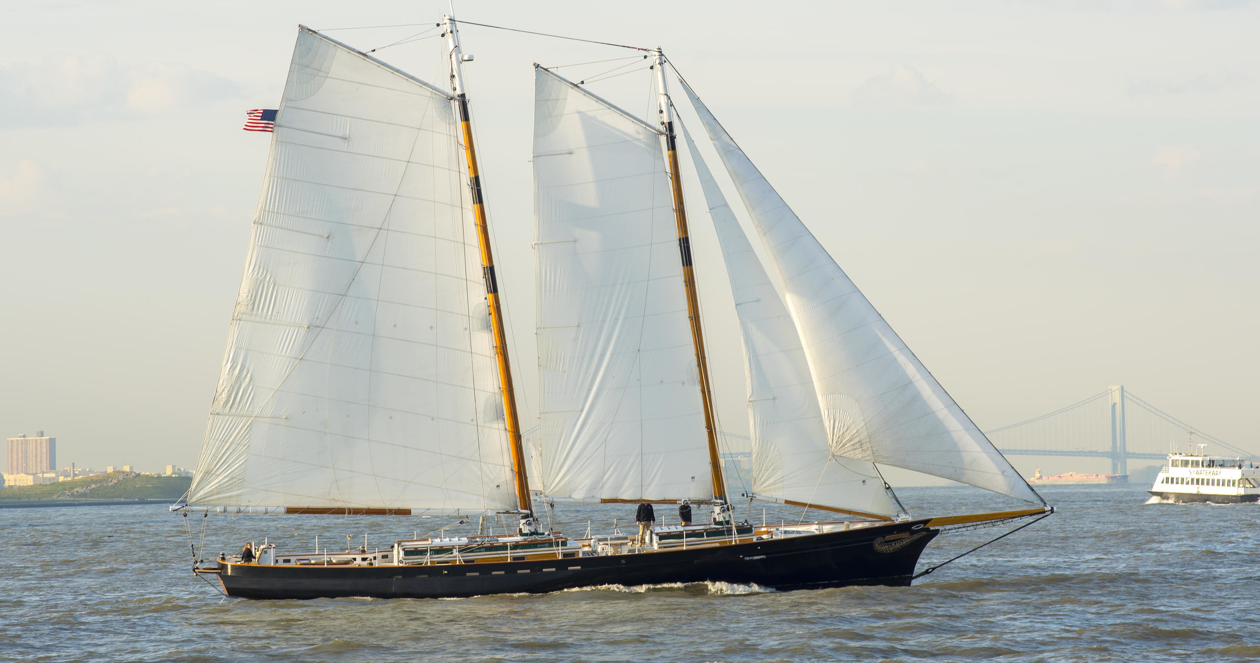 A two-masted sailboat on calm water with a bridge in the background.
