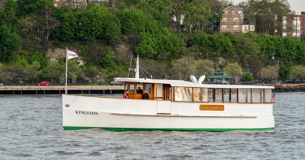 White tour boat named Kingston on a river with trees and buildings in the background.