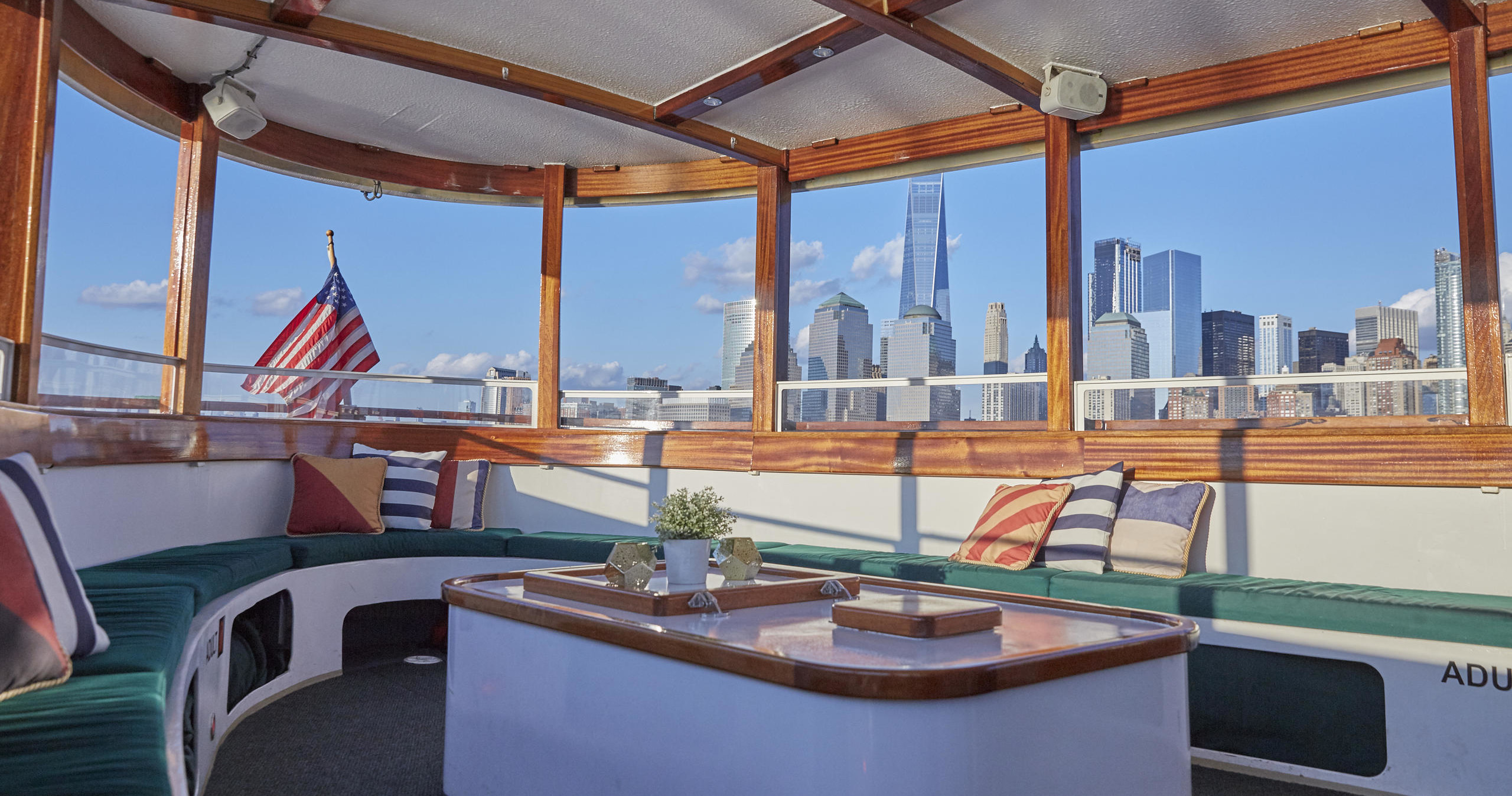 Boat interior with city skyline view through windows, featuring seating and an American flag.