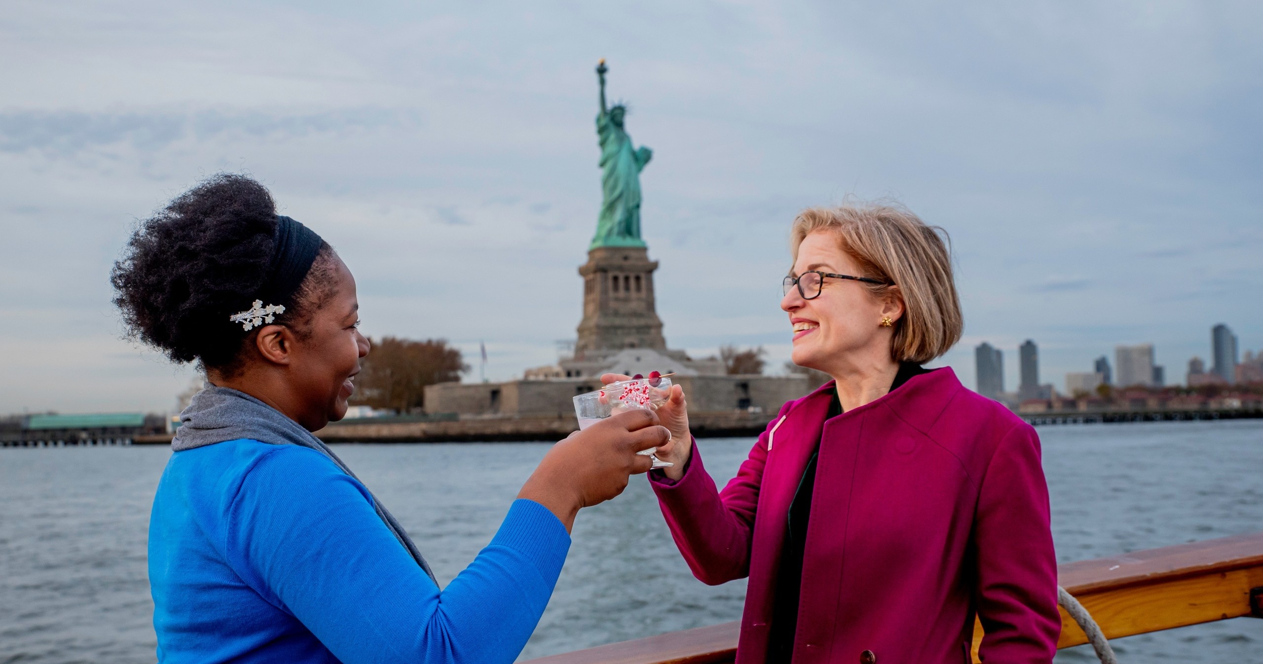 Two people toast on a boat with the Statue of Liberty in the background.