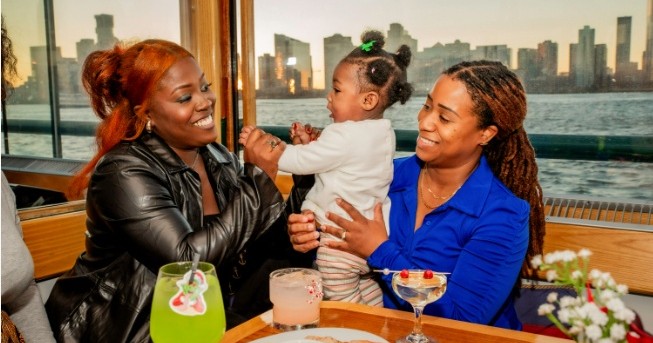 Two women smiling at a baby on a boat with a city skyline in the background.