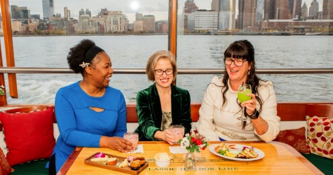 Three women laughing and having drinks on a boat with city skyline in the background.