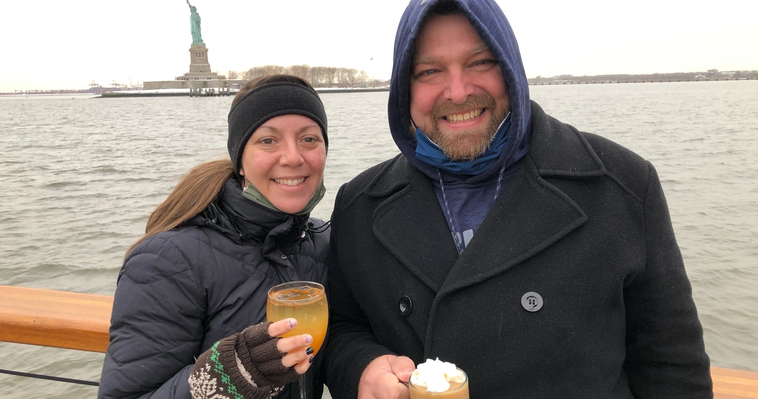 Two people in winter clothing holding drinks with the Statue of Liberty in the background.