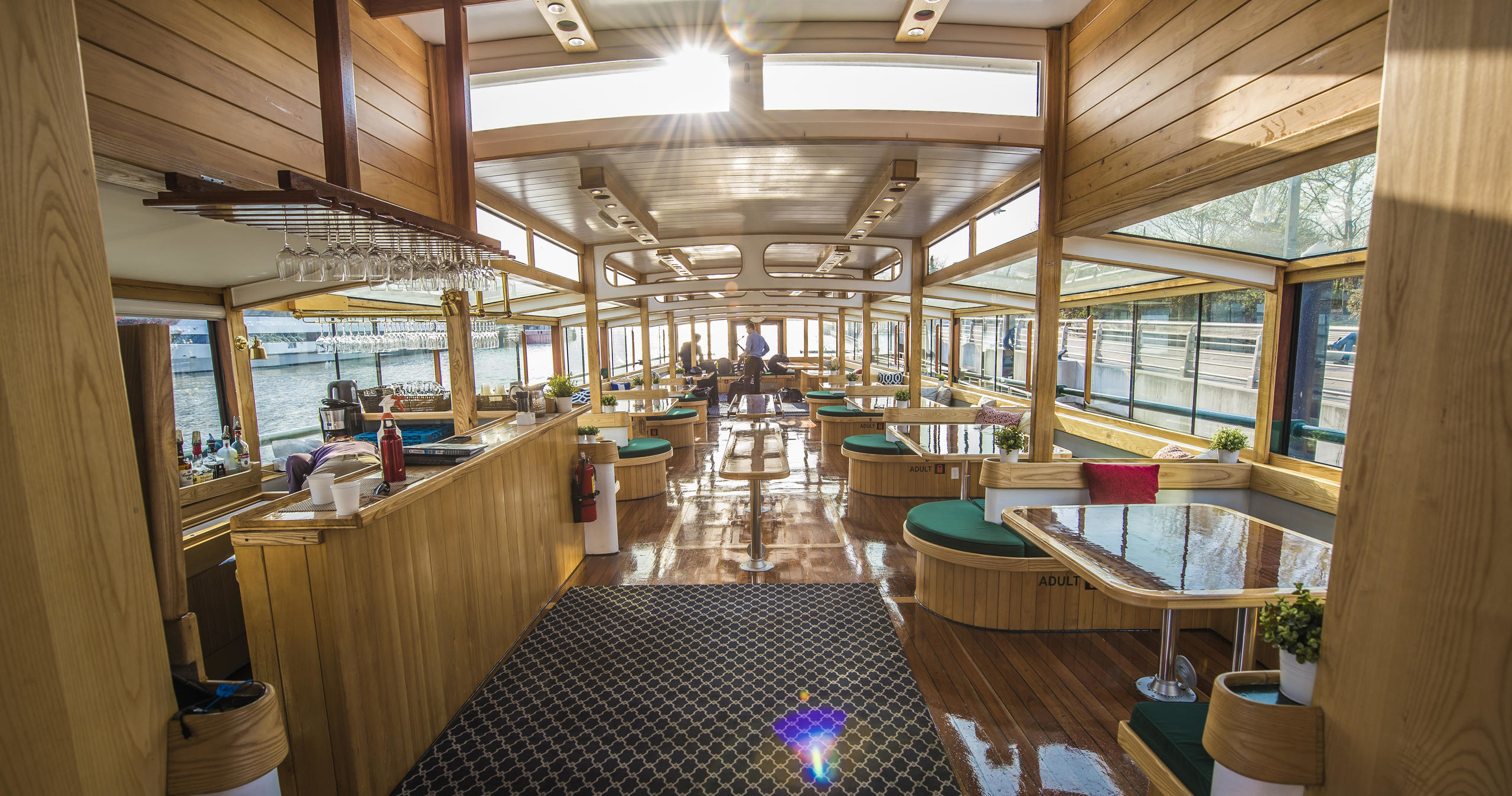 Luxurious boat interior with wood paneling, large windows, sunlit seating, and a bar area.