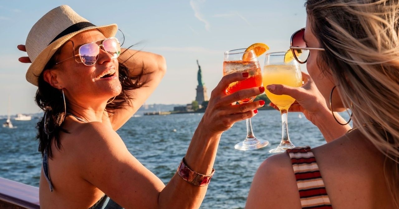 Two women clinking glasses on a boat with Statue of Liberty in the background.
