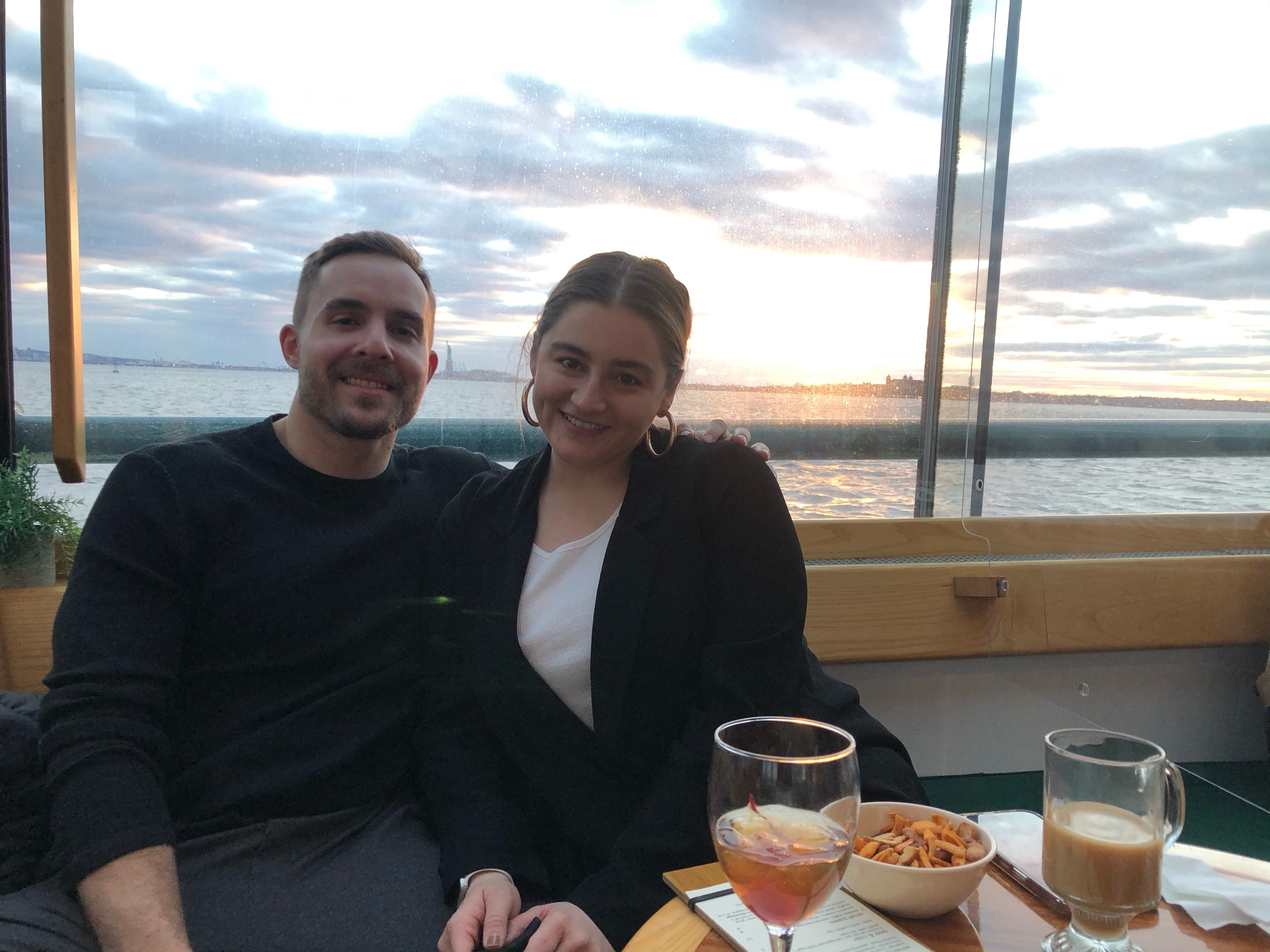Smiling couple seated by a window with snacks and drinks, sunset view over water in background.